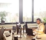 A businessman sits at a desk using multiple computers and a headset in a well-lit modern office.